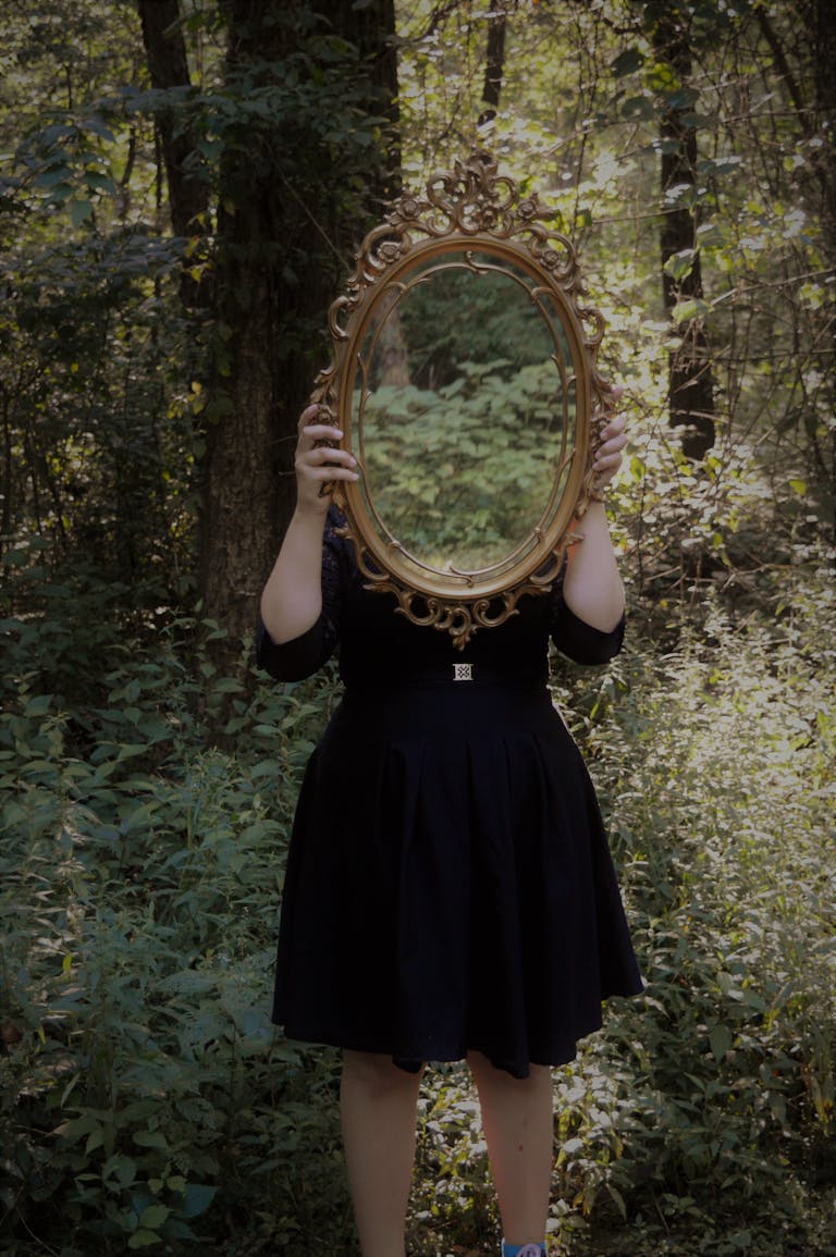 Η σκιά, το πέπλο και η αλήθεια 1 A woman holds an ornate mirror in a forest setting, reflecting nature.